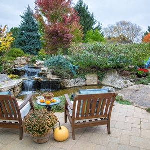 Two wooden chairs face a pond and waterfall in a colorful autumn garden with pumpkins.