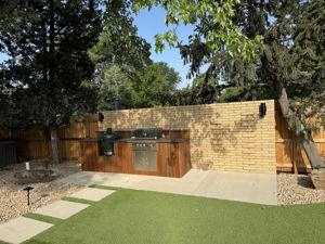 Outdoor kitchen with a grill, wooden cabinets, and a stone wall. Green grass and trees surround it.