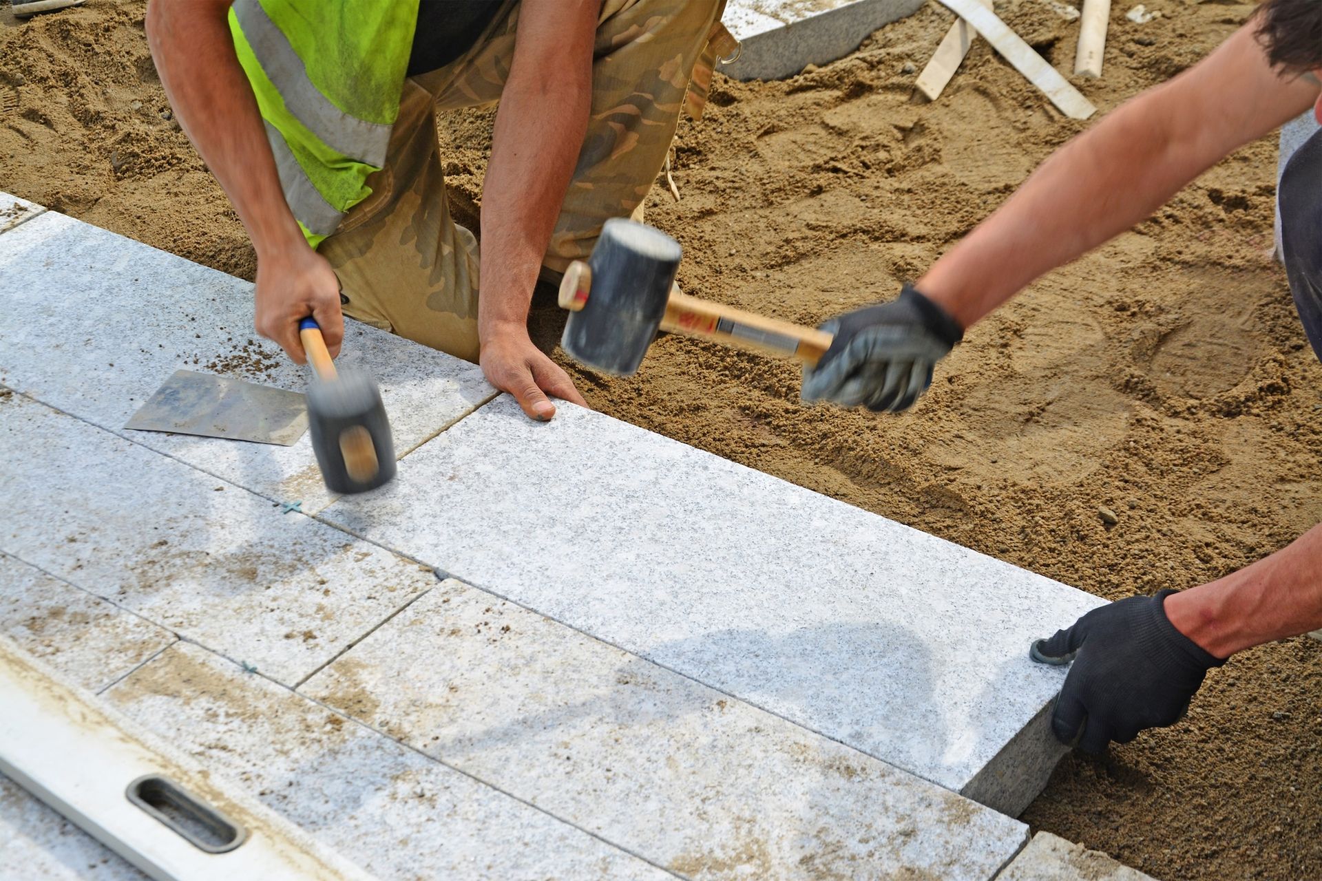 Two workers laying stone pavers, one uses a mallet, other a trowel, outdoors on dirt.