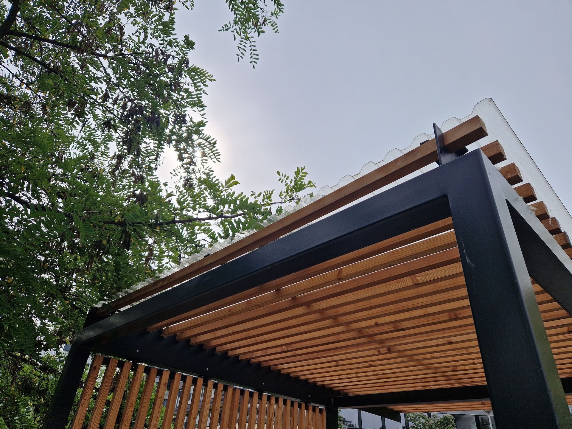 Black and wood pergola with horizontal slats. Trees and sky visible above.