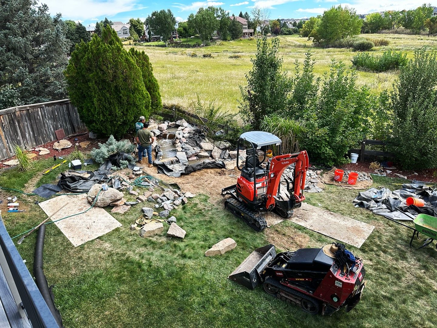 Backyard construction site; mini-excavator and skid-steer; person working amidst rocks and debris.