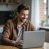 Man smiling while typing on a laptop at a table near a window
