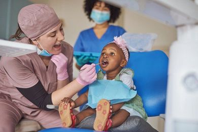 Dentist examining a young Black child's teeth; assistant in background. Clinic setting; pink, blue, and white colors.