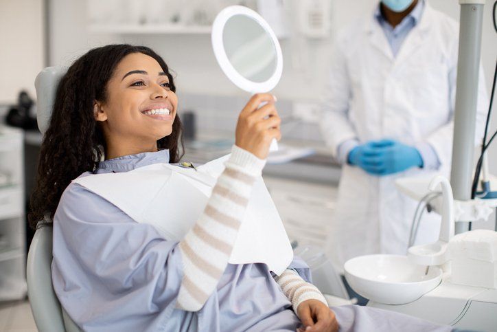 Smiling woman at the dentist, looking at her teeth in a mirror, with a dentist in the background.