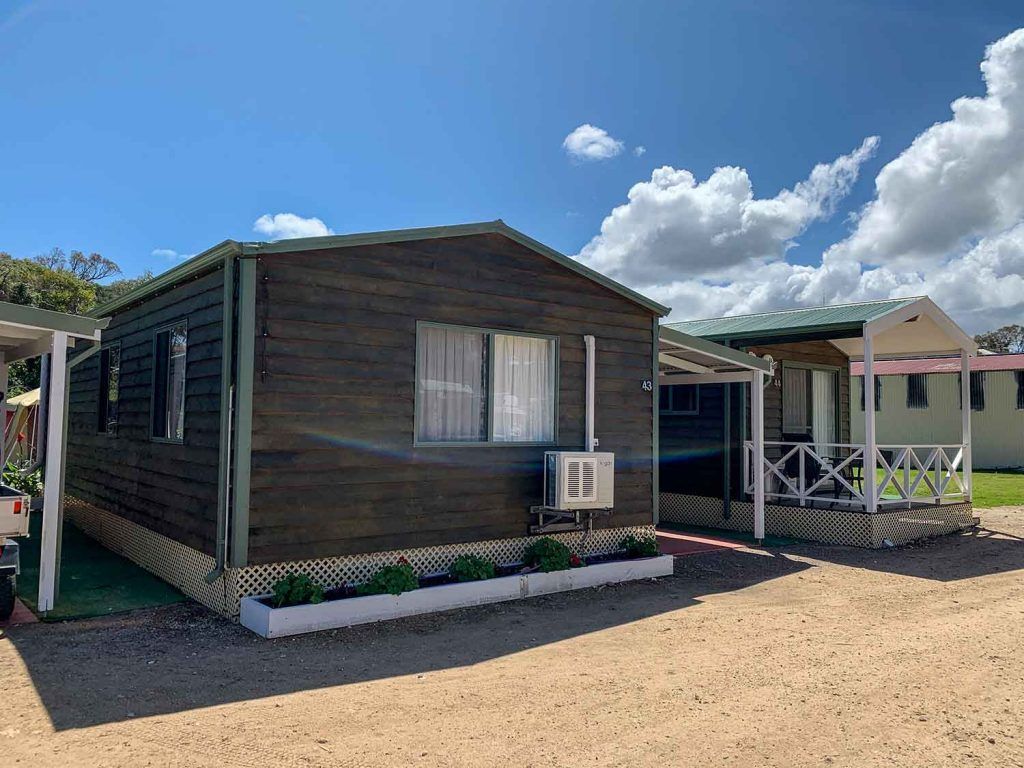 Small dark wooden cabin with a white porch and air conditioner under a blue sky at a campsite