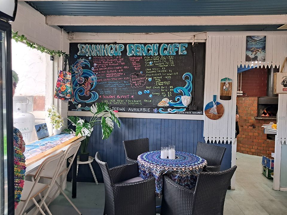 Cozy beach café patio with chalkboard menu, wicker chairs, blue tablecloths, and plants.