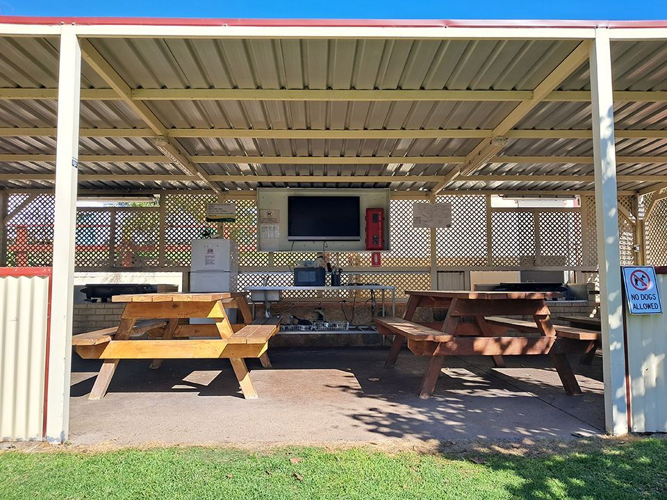 Covered picnic area with two wooden picnic tables and a wall-mounted TV under a metal roof