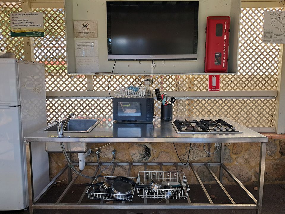 Commercial kitchen prep area with stainless steel table, sink, stove, fridge, and wall-mounted TV.