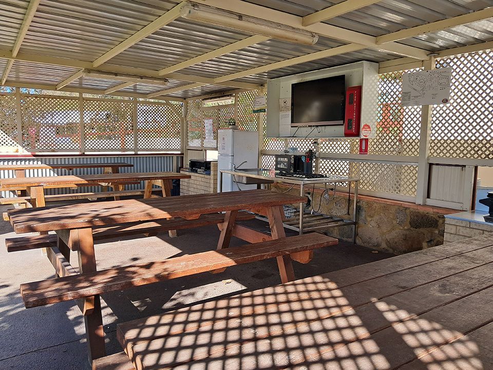 Covered picnic area with wooden tables, lattice walls, and a TV mounted on the wall in sunlight.
