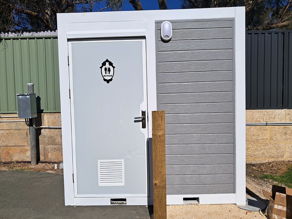 White and gray outdoor utility shed with a door, vent, and wall-mounted light on a sunny day