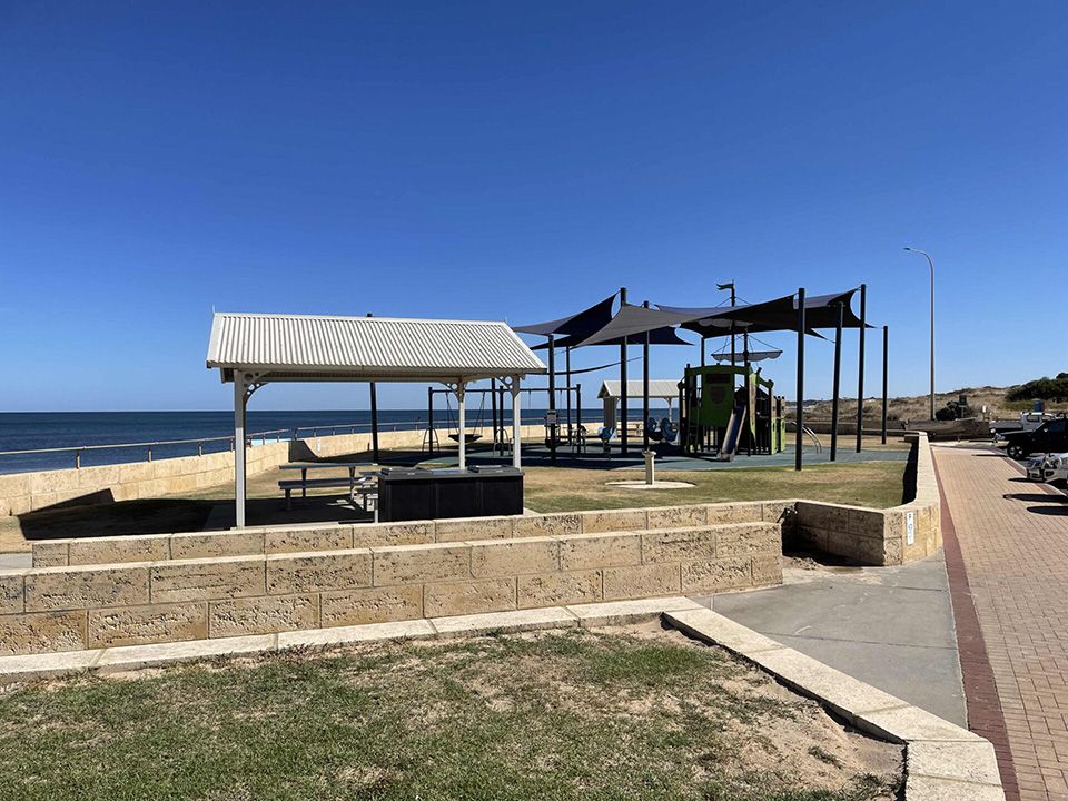 Beachside playground with shade structures, benches, and a walkway by the ocean