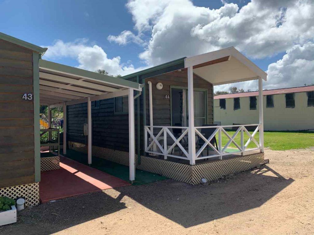 Small dark cabin with white porch railings and red awning in a sunny mobile home park.