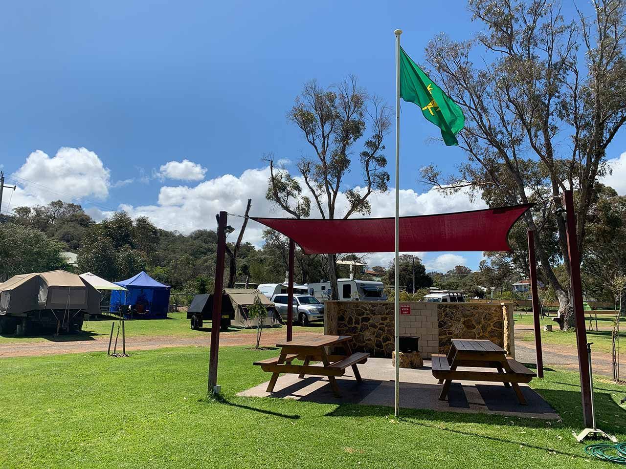 Outdoor campsite with picnic tables under a red canopy and green flag on a sunny grassy field.