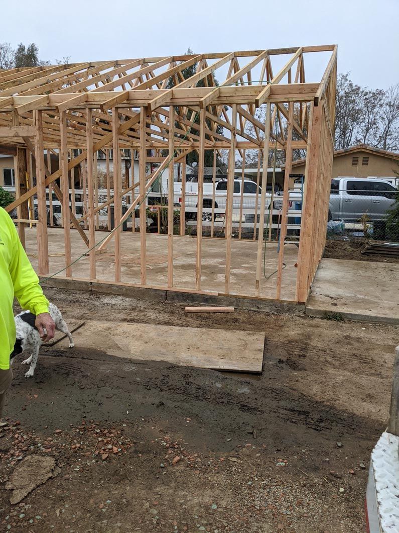 Partially built wooden house framing on a muddy construction site, with a worker in neon yellow at left.