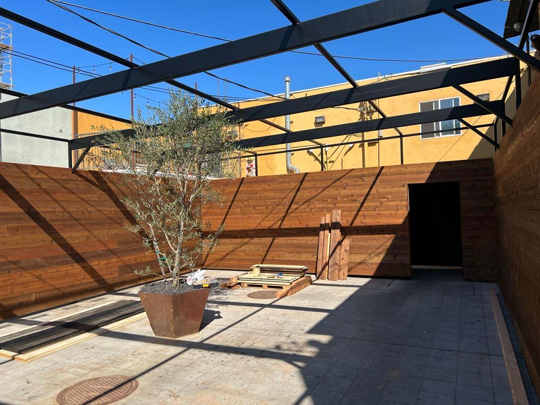 Sunlit rooftop patio with wooden walls, potted tree, and a shaded seating area under a steel frame.