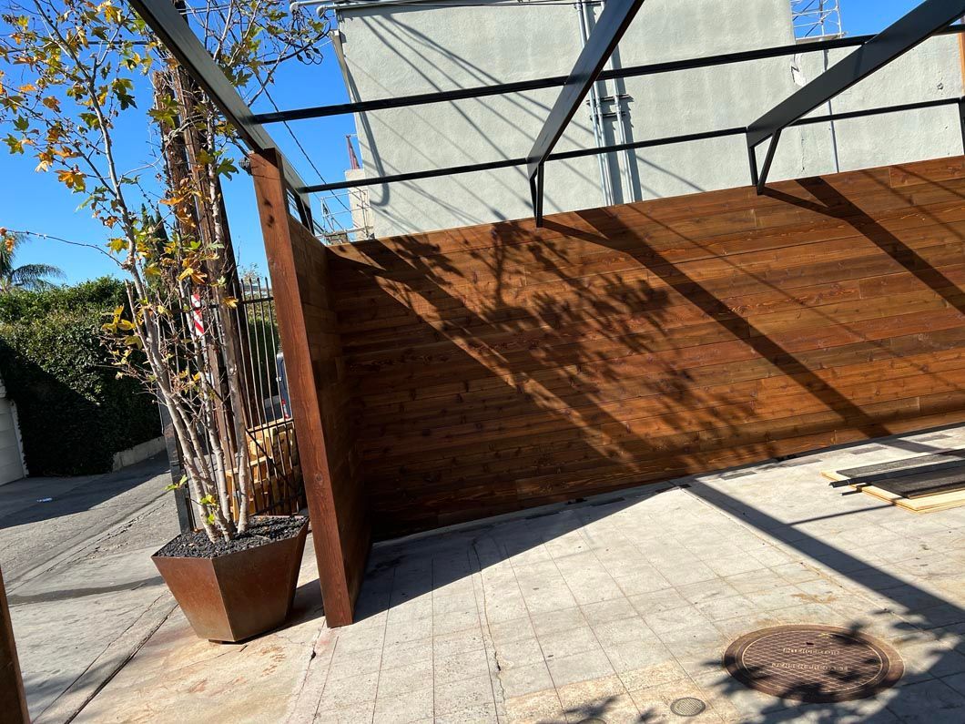 Outdoor patio with wooden privacy wall, potted tree, and sunlight casting shadows on concrete.