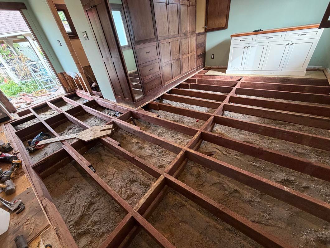Exposed wooden floor joists in a room under renovation, with cabinets and a window in the background