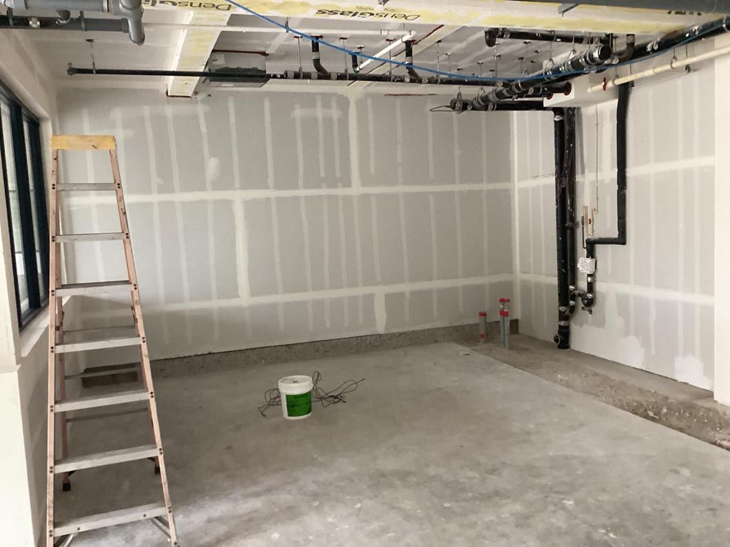 Empty unfinished room with drywall, ladder, and paint bucket on a concrete floor.