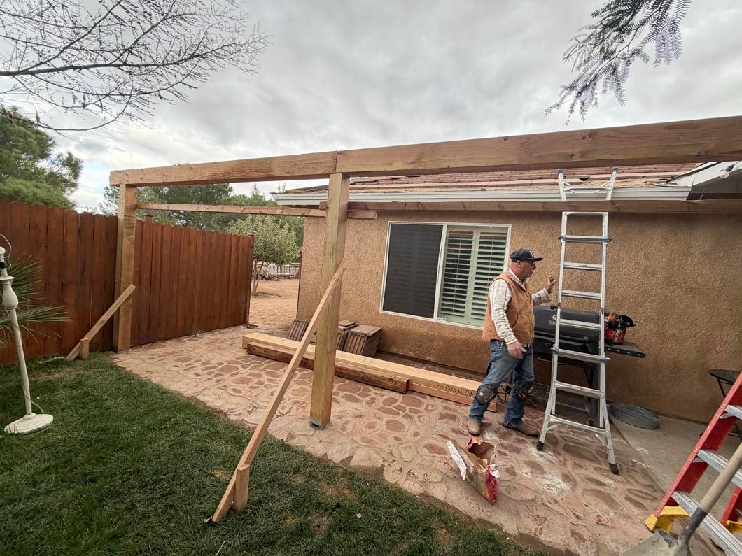 Man building a wooden patio pergola in a backyard, with ladder, tools, and fence nearby.