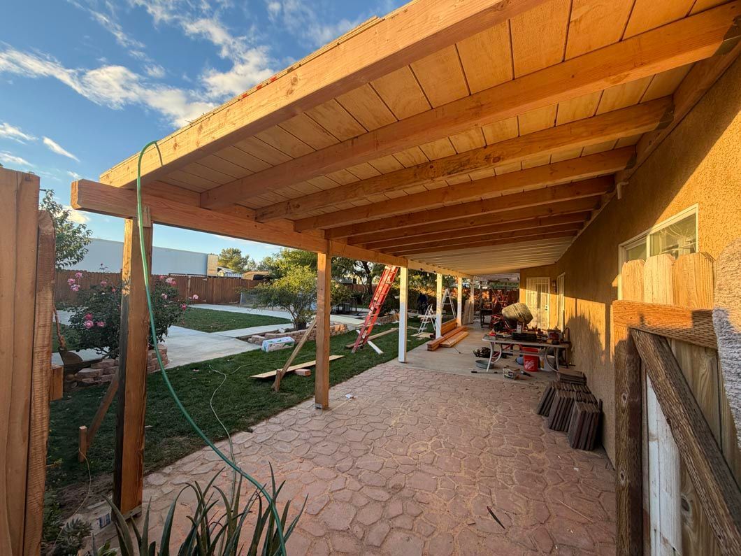 Covered patio with wooden beams, stone floor, and garden beside a brick house at sunset
