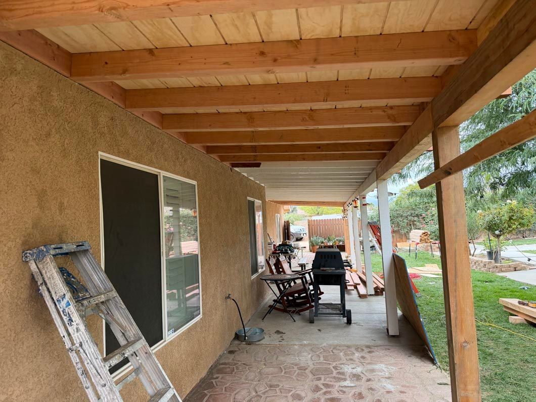 Covered porch with wood ceiling, ladder, chairs, and stucco wall beside a walkway and yard