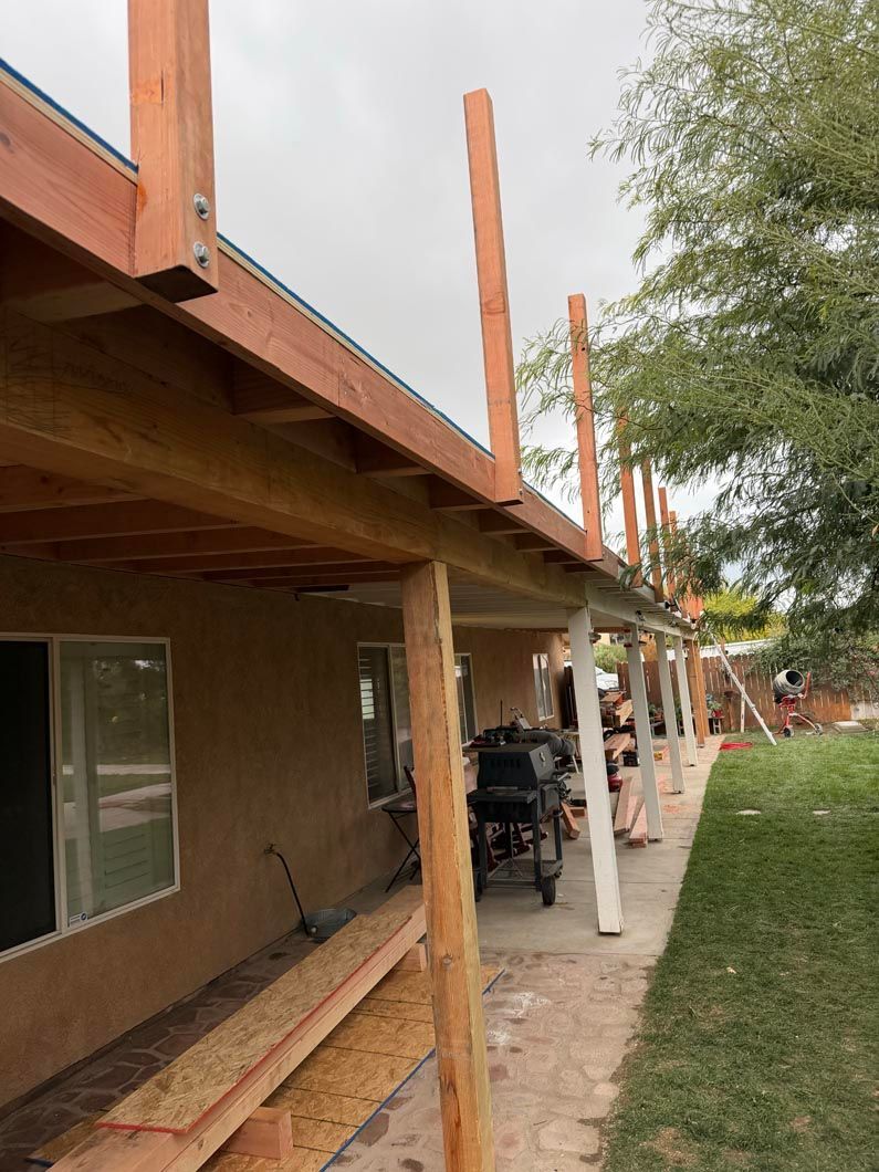 Outdoor building under construction with unfinished roof, wooden supports, and construction materials along a dirt path.