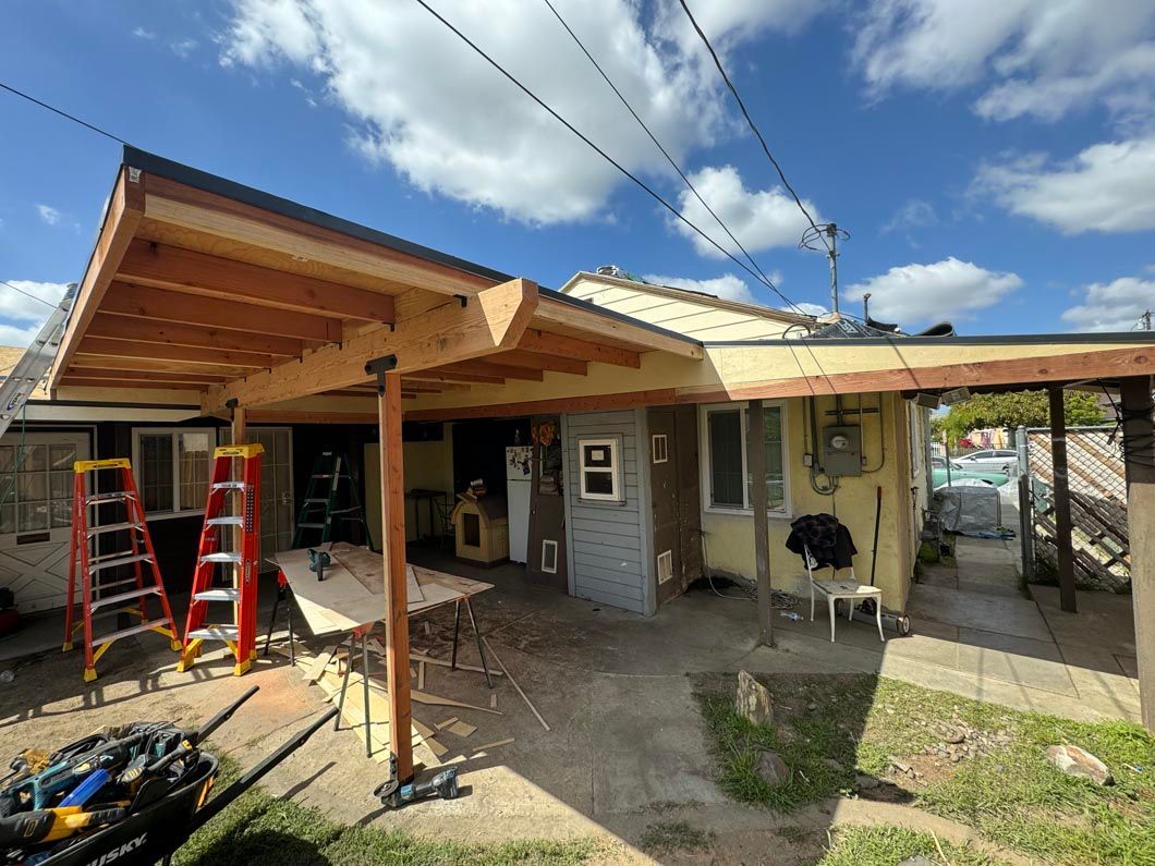 Partially constructed patio roof with ladders and tools beside a small house under a blue sky