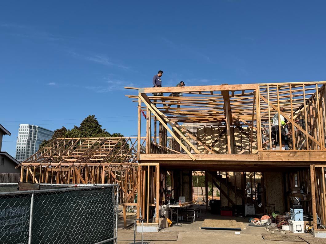 House under construction with exposed wooden framing and workers on the roof under a clear blue sky