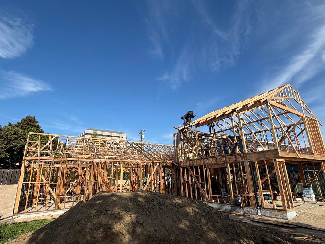 Wooden house frame under construction on a sunny day, with blue sky and a dirt mound in front