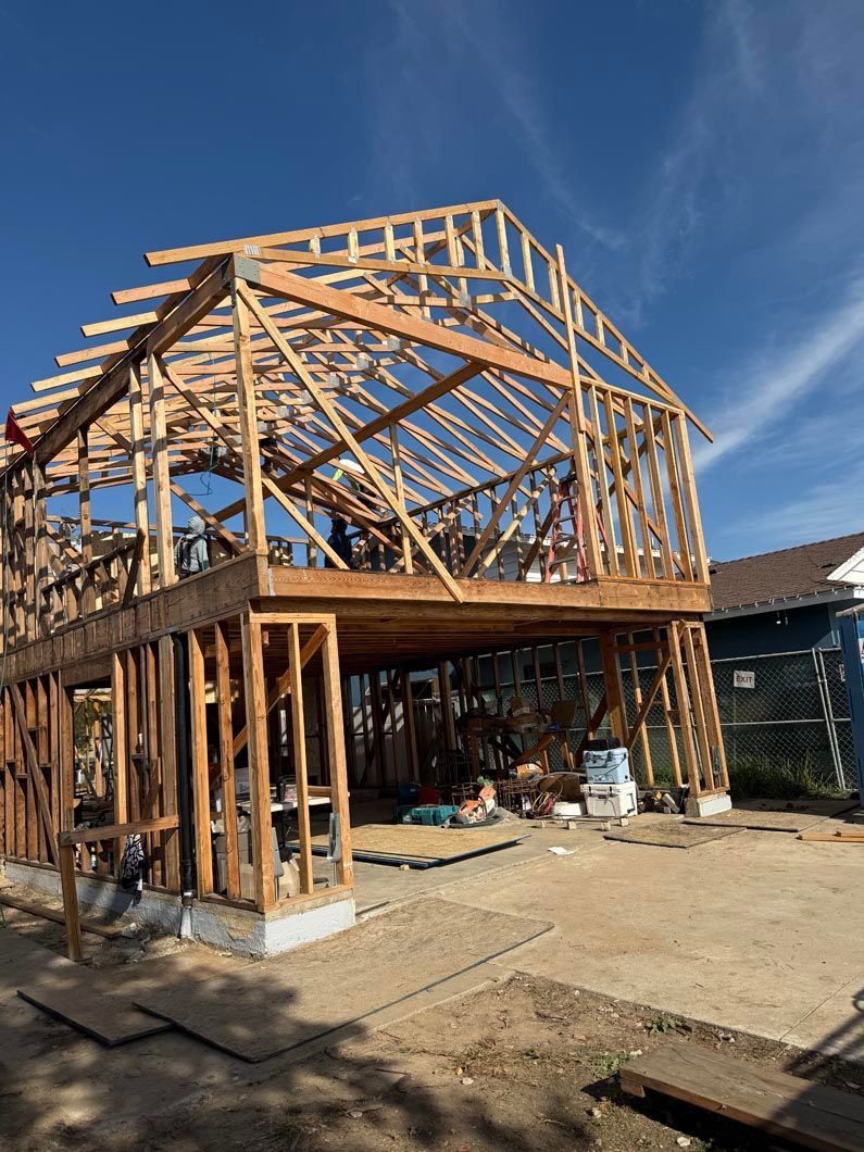 Wood-framed house under construction with exposed beams and roof trusses on a sunny day