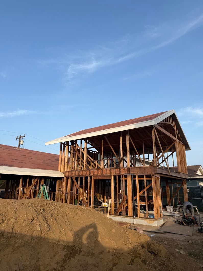 Partially built wooden house with red roof under a clear blue sky, surrounded by dirt and construction materials