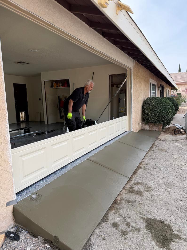 Worker smoothing fresh concrete walkway beside a house with a large open window.