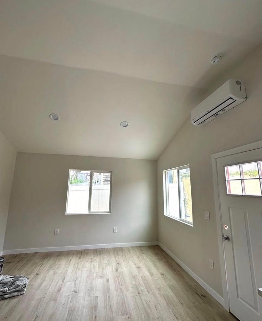 Empty beige living room with light wood floors, two windows, a front door, and a wall-mounted AC unit
