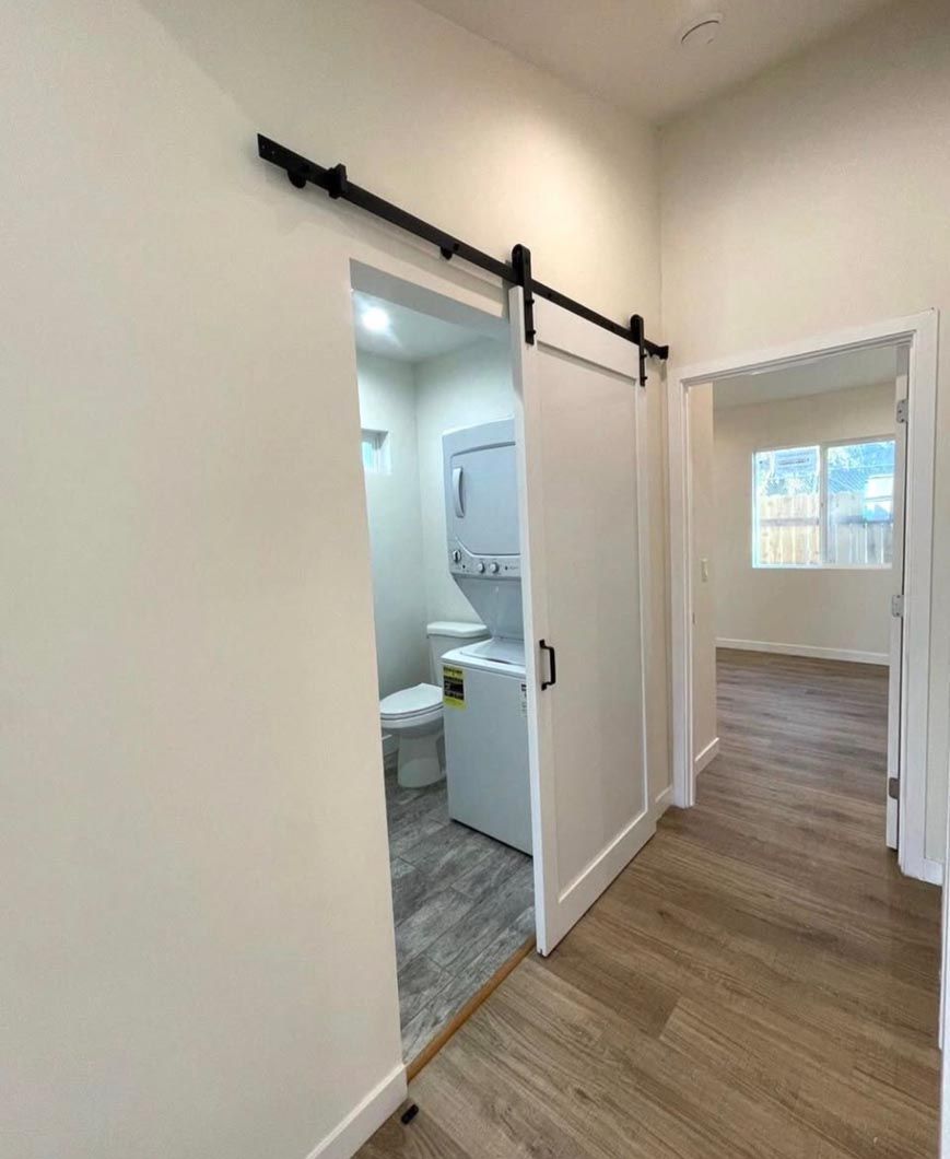 Open doorway to a small bathroom with gray tile, beside a bright room with wood floors and a window.