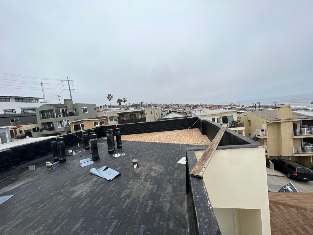 Rooftop with dark tar surface, vents, and white walls overlooking a coastal neighborhood under a cloudy sky
