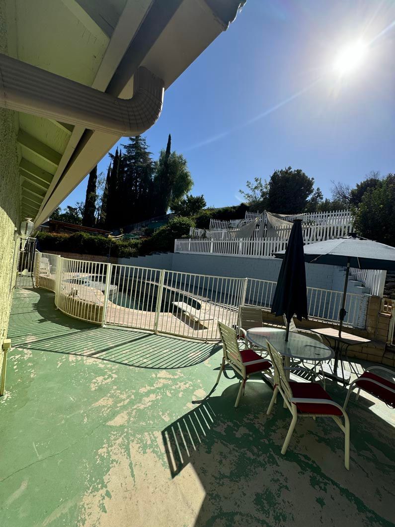 Sunny patio with chairs and table under a white canopy, overlooking a fenced hillside yard.