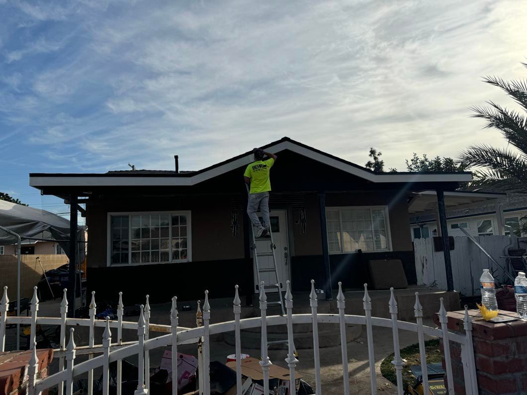Person in a neon vest on a ladder painting a house exterior behind a white fence.