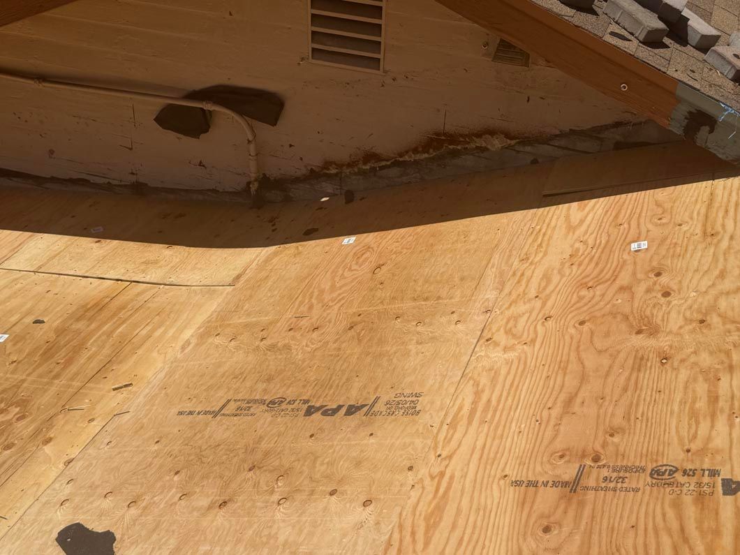 Close-up of plywood sheathing beneath a roof overhang, with tan wood and metal framing visible.