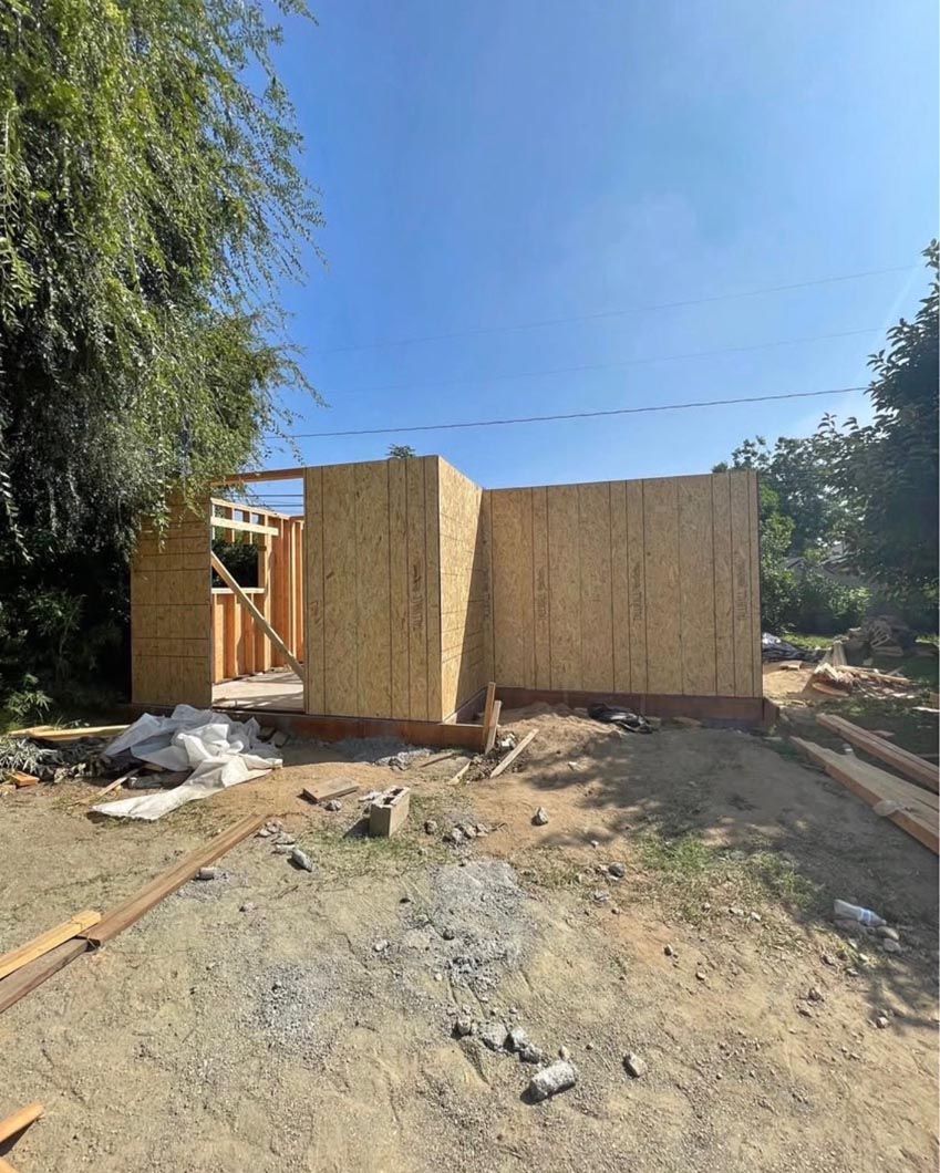 Partially built wooden shed in a dirt yard, with trees and a clear blue sky behind it