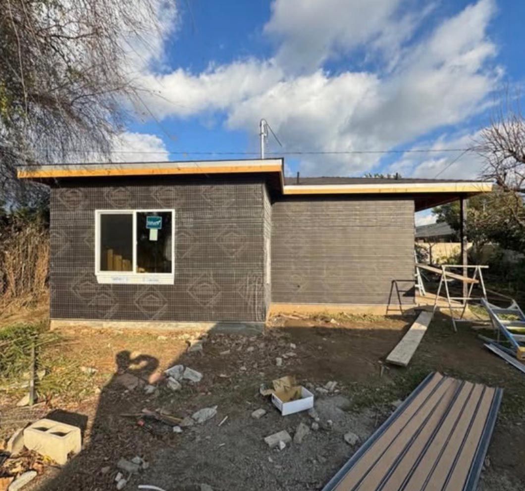 Small modern gray house exterior under construction, with tools and lumber in the yard and a blue sky overhead.