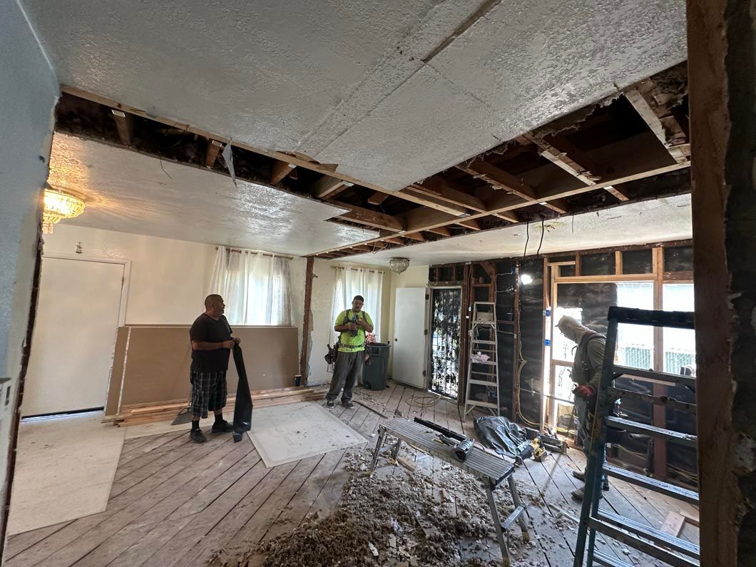 Workers renovating a room with exposed ceiling joists, drywall, and construction debris on the floor.