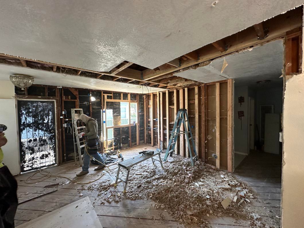 Interior under renovation with exposed studs, debris on the floor, and workers near windows