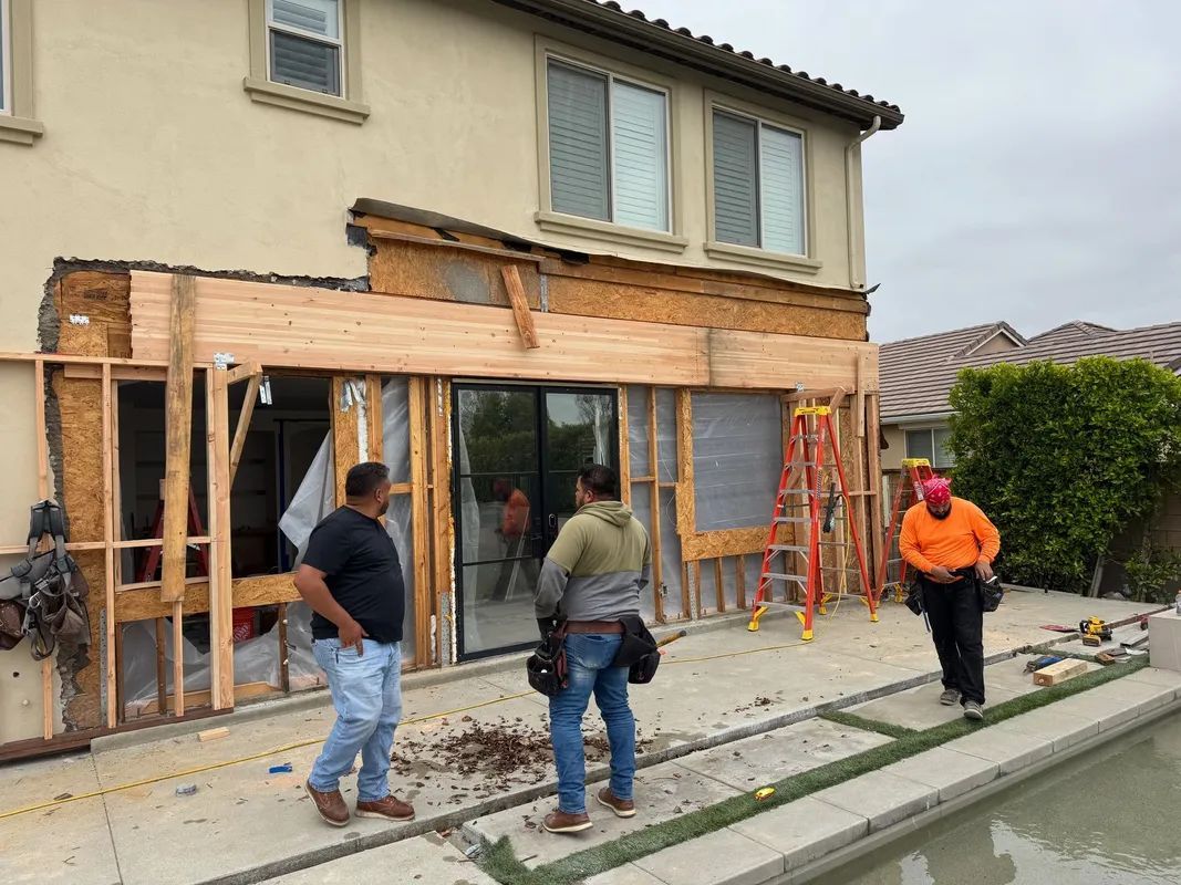 Workers renovating a house exterior with wood framing, ladders, and scaffolding beside a canal