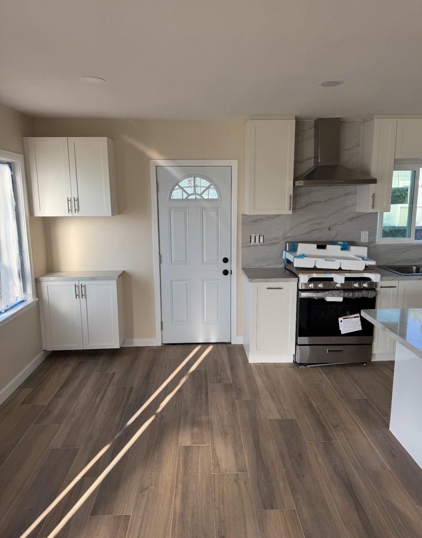 Empty kitchen with white cabinets, stainless stove, wood floor, and sunlight streaming through a window.