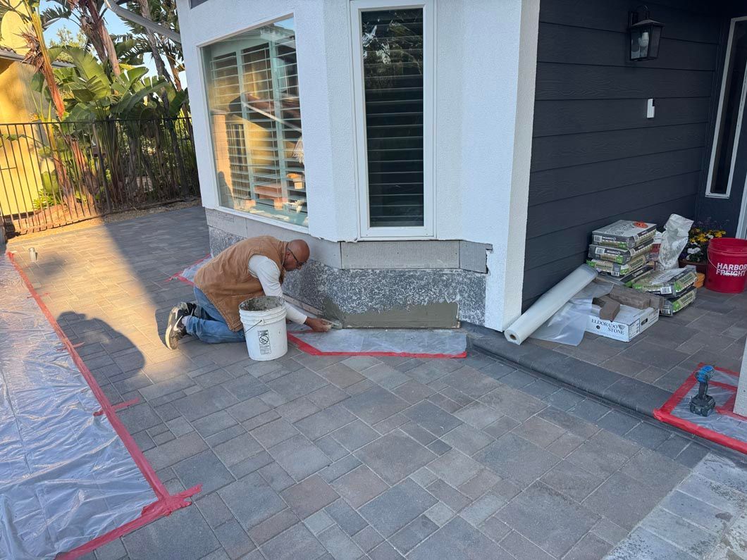 Person kneeling on a patio, applying material near a house window and door beside buckets and supplies.
