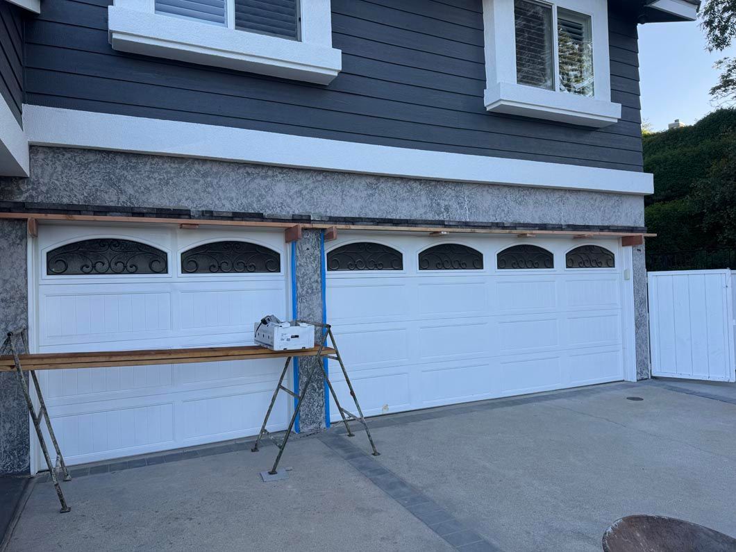 Two garage doors with white and gray siding; sawhorses and boards set up on the driveway.