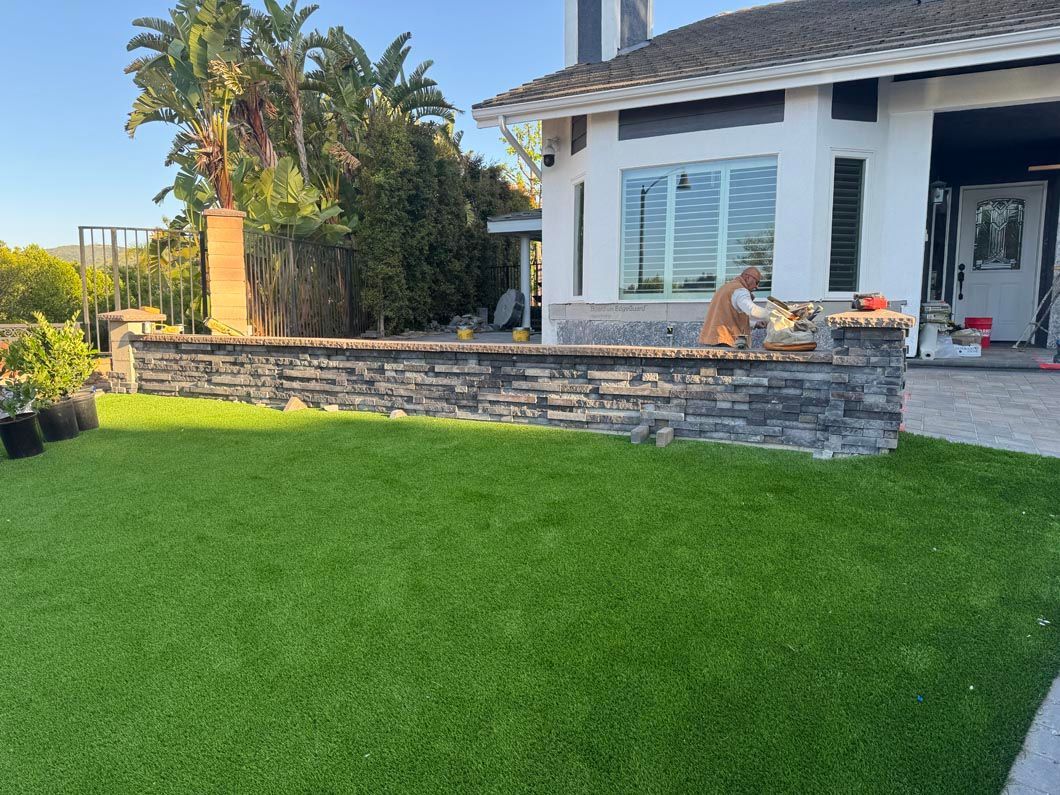 Backyard patio with green lawn, stone planter wall, and a white house in the background