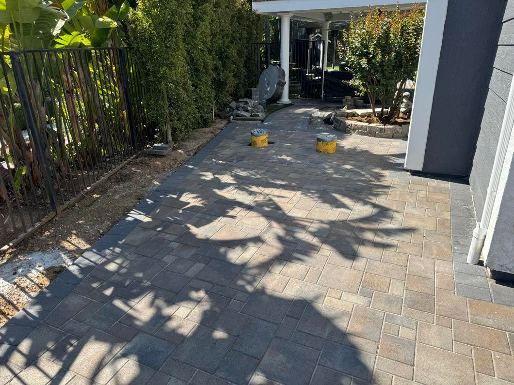 Backyard patio with pavers, hedges, and scattered yellow toys in bright sunlight