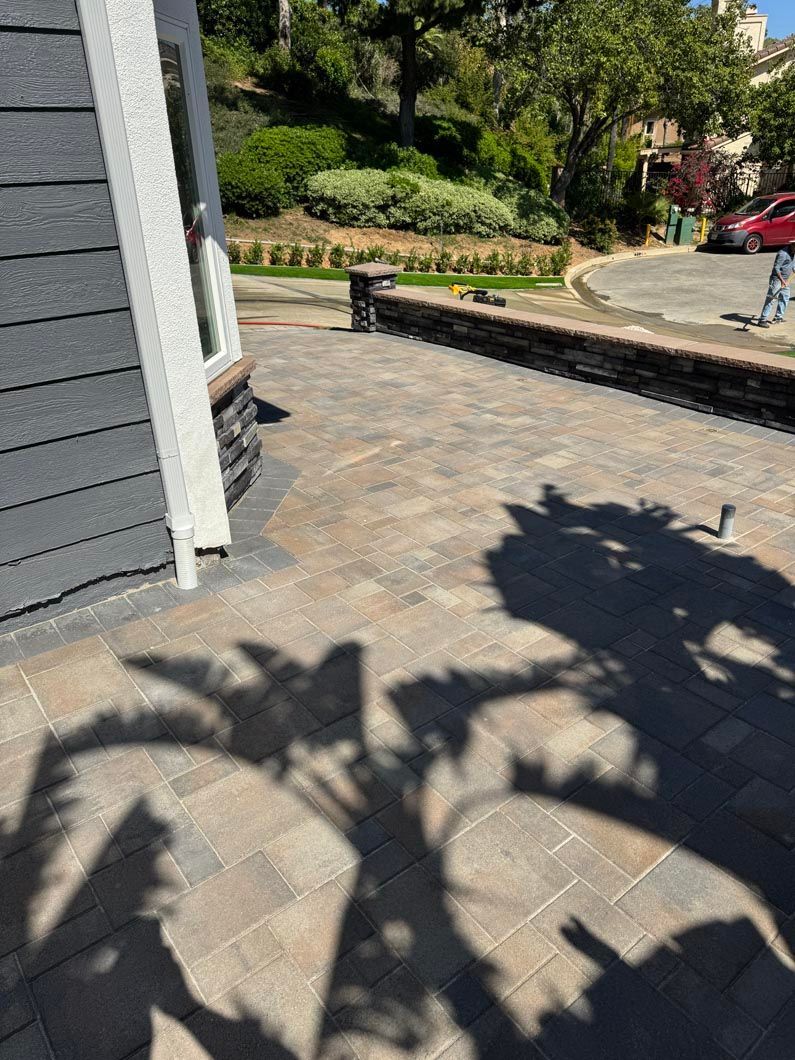 Sunlit stone patio beside a house, with a garden wall, trees, and shadows in the foreground.