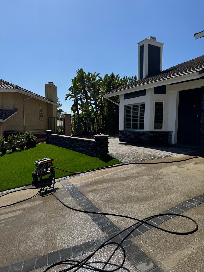Sunlit suburban driveway beside a white house with black trim, green lawn, and a pressure washer hose in front.